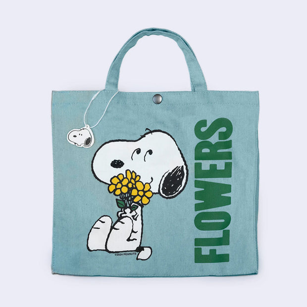 Light blue fabric tote bag with a short purse-like handle and a snap closure. On the front is a graphic of Snoopy, sitting and smiling while holding a yellow bouquet of flowers. Large green text reads "Flowers" along the side.