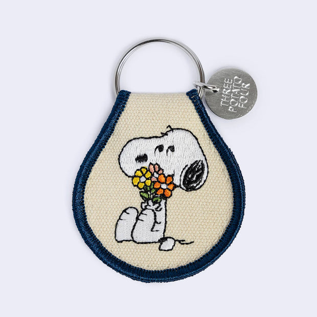 Embroidered keychain, cream with a dark blue border and featuring and illustration of Snoopy sitting and holding a bouquet of flowers. Fabric is attached to a keyring and a small silver disc that reads "three potato four"