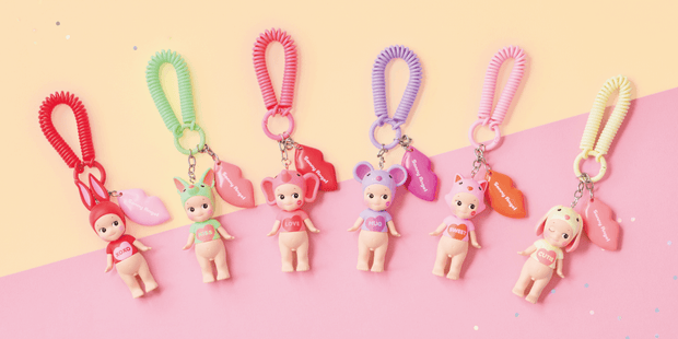 Promo photo featuring 6 different Sonny Angel figurines, all nude on the bottom but wearing t-shirt of Valentine's themed colors that say sentiments such as "hug", "love", "xoxo", etc. They all wear hood hats that are shaped like animal heads matching their t-shirt colors. Each are attached to a keychain with a lip shaped charm.