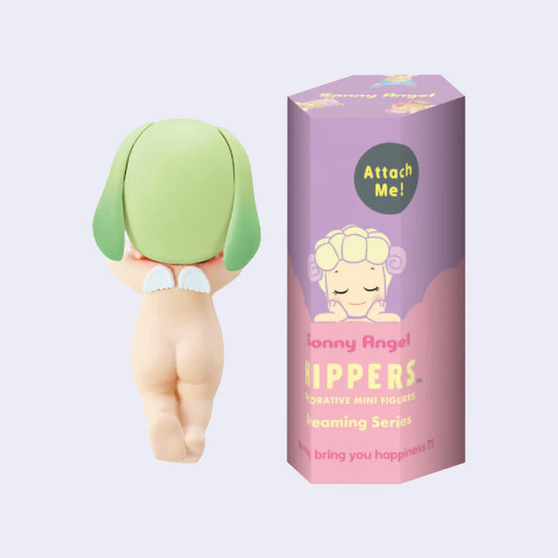 A small Kewpie baby figure, facing away so only their nude body is showing, their legs slightly crossed with a small pair of angel wings on their back. They wear a green lop eared bunny hood hat. Next to them is a pink and purple color display box with an illustration of a smiling Kewpie baby with a sheep hood hat resting its arms flat. 