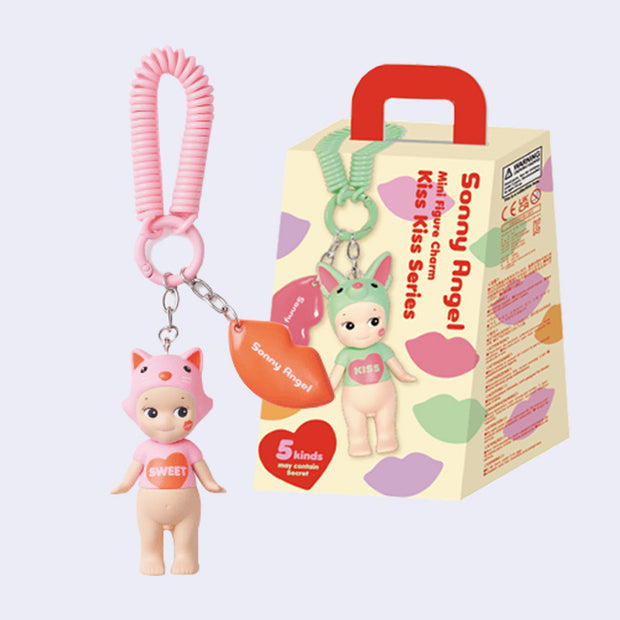 Mini figure of a Kewpie baby nude aside from a pink t-shirt that says "sweet" on it in a heart and a pink hood hat in the shape of a cat. It is attached to a pink rubber chain, alongside a red charm of lips that reads "Sonny Angel". Positioned next to its product packaging. 