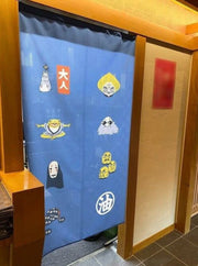 View of a blue tapestry Spirited Away characters and illustrated iconography on it hanging as a partition between 2 rooms.