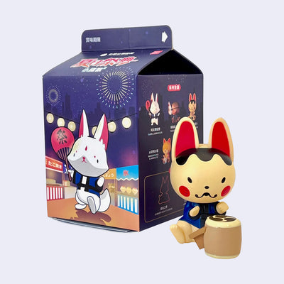 Small figure of a Japanese style traditionally designed fox, with large red ears and rosy cheeks. It sits wearing a blue tied shirt and bangs on a drum.