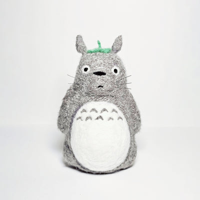 Needle felted wool sculpture of Totoro, standing with a curious expression on his face and arms by his side. He has a leaf atop his head and stiff whiskers.