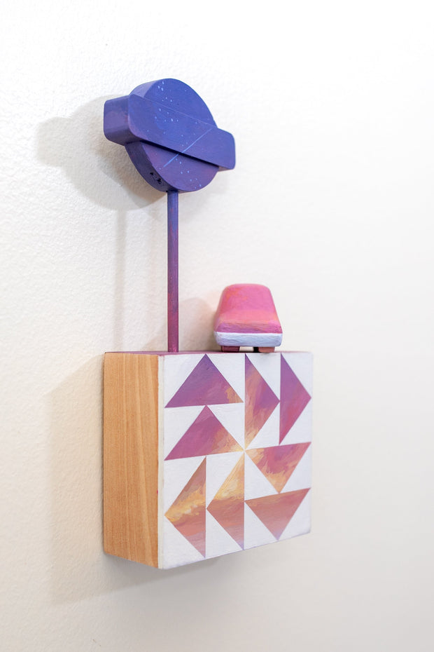 Small square wood panel painted like a pink sunset with white patterning akin to a quilt tile design. Atop of it are 2 carved wooden sculptures: one of a pink retro looking car and the other of a dark purple signage attached to a long pole.