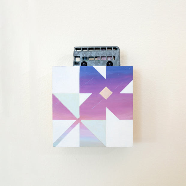 Small square wood panel painted like a purple sunset with light blue and white patterning overlayed on top, making a flower tile design. Atop of it is a small sculpture of a double decker bus, painted to look very old.