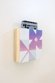 Small square wood panel painted like a purple sunset with light blue and white patterning overlayed on top, making a flower tile design. Atop of it is a small sculpture of a double decker bus, painted to look very old.