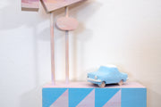 Small square wood panel painted like an light pink sunset with blue patterning overlayed on top, akin to a quilt tile design. Atop of it are carved wooden sculptures: a blue retro style car and several large sunset colored Americana style signs, attached to 2 tall poles.