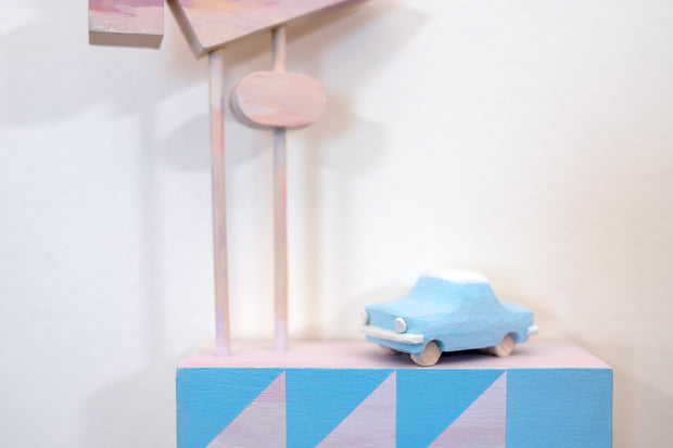 Small square wood panel painted like an light pink sunset with blue patterning overlayed on top, akin to a quilt tile design. Atop of it are carved wooden sculptures: a blue retro style car and several large sunset colored Americana style signs, attached to 2 tall poles.