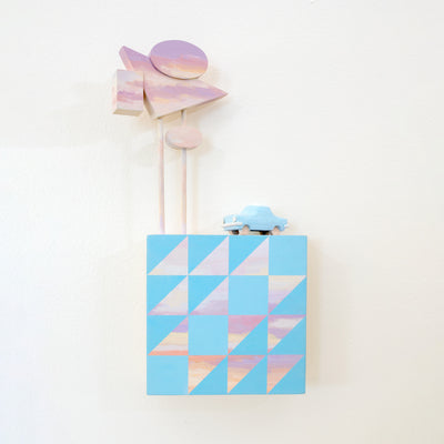 Small square wood panel painted like an light pink sunset with blue patterning overlayed on top, akin to a quilt tile design. Atop of it are carved wooden sculptures: a blue retro style car and several large sunset colored Americana style signs, attached to 2 tall poles.