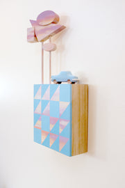 Small square wood panel painted like an light pink sunset with blue patterning overlayed on top, akin to a quilt tile design. Atop of it are carved wooden sculptures: a blue retro style car and several large sunset colored Americana style signs, attached to 2 tall poles.