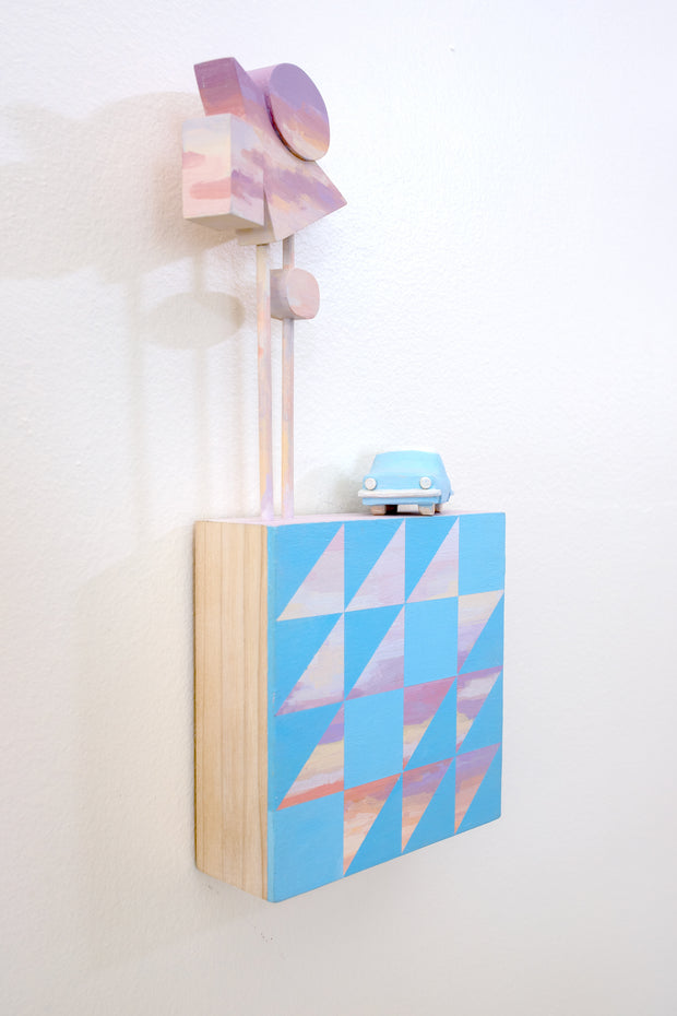 Small square wood panel painted like an light pink sunset with blue patterning overlayed on top, akin to a quilt tile design. Atop of it are carved wooden sculptures: a blue retro style car and several large sunset colored Americana style signs, attached to 2 tall poles.