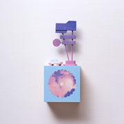 Small square wood panel with a purple and pink sunset shown within a flower outline, the rest of the panel is painted light blue. Atop of it are carved wooden sculptures: a small pink colored retro car and a several large signs attached to 2 poles.