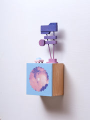 Small square wood panel with a purple and pink sunset shown within a flower outline, the rest of the panel is painted light blue. Atop of it are carved wooden sculptures: a small pink colored retro car and a several large signs attached to 2 poles.