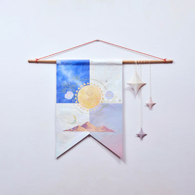 Painted canvas banner hanging from a wooden dowel. The banner is divided into 4 parts: blue night sky, white, pink sunset and light colored clouds with a painted sun and flowers in the middle of all 4 and a painted mountain range along the bottom. 