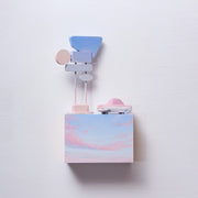 Small rectangular wood panel painted like a light blue sky with a pink sunset. Atop of it is a small wooden car and a  series of large Americana style signs attached to the same pole.