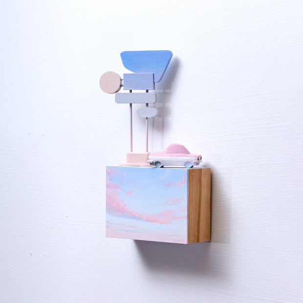 Small rectangular wood panel painted like a light blue sky with a pink sunset. Atop of it is a small wooden car and a  series of large Americana style signs attached to the same pole.