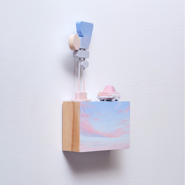 Small rectangular wood panel painted like a light blue sky with a pink sunset. Atop of it is a small wooden car and a  series of large Americana style signs attached to the same pole.