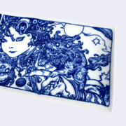 Rectangular shaped white porcelain plate with dark blue line art illustrations, depicting a woman with miniature lion like creatures on her shoulder and under her face. She looks at the viewer with a serious expression. To the left are illustrated panels of a person riding a dragon and another person creating a laser with their fingers. Close up.