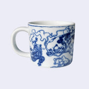 White porcelain mug with a dark blue line art illustration wrapped around it. A small man with a big nose fights a large koi fish with smoke lingering behind it.