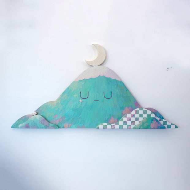 Die cut wooden panel with a painting of a teal colored mountain. It has a simple closed eye facial expression and smaller mountains are layered in front of it, one with a white checkered pattern. A crescent moon rests atop the mountain.