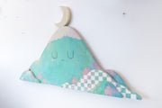 Die cut wooden panel with a painting of a teal colored mountain. It has a simple closed eye facial expression and smaller mountains are layered in front of it, one with a white checkered pattern. A crescent moon rests atop the mountain.