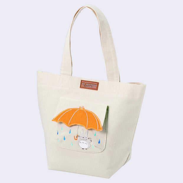 Cream colored handle bag with a front pocket featuring an embroidery of Totoro holding a large yellow umbrella with blue raindrops around. A pleather sewn plaque in the center of the bag reads 'My Neighbor Totoro"