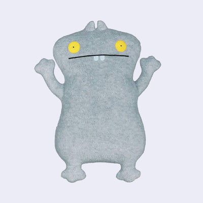 Fluffy gray plush doll of a mostly flat standing monster with yellow eyes and a straight lined, wide mouth with 2 small teeth showing.