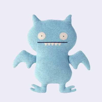 Light blue plush monster doll, mostly flat in shape, with sewn on eyes and a straight mouth smile with 5 sharp pointed teeth.