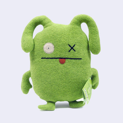 Green plush doll of an ugly yet very cute monster with floppy ears like a rabbit, one x eye and another circular eye. It has a straight mouth with a small tongue sticking out.