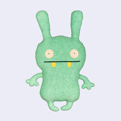 Plush doll, mostly flat in shape, of a bunny like character with wide set circular eyes and a straight lined mouth with 2 yellow round teeth peeking out.