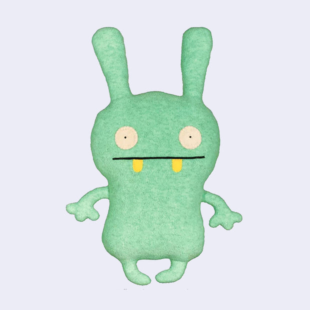 Plush doll, mostly flat in shape, of a bunny like character with wide set circular eyes and a straight lined mouth with 2 yellow round teeth peeking out.