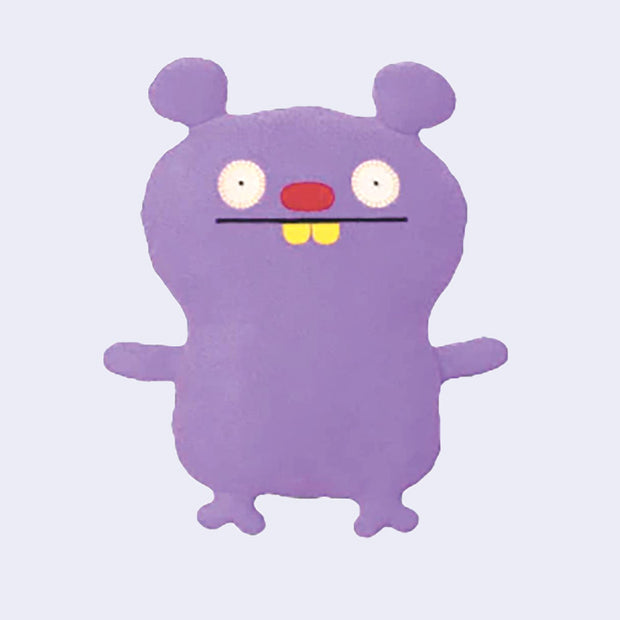 Plush doll of a purple monster creature with small round ears like a bear and a mostly flat body. It has wide set circular eyes, a red oval shaped nose and 2 yellow buck teeth.