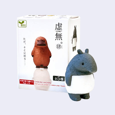 Small plastic figure of a tapir, grey with a white belly standing on its hind legs with its arms to its side and looking off with a blank expression. Its product packaging is behind.