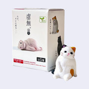 Small figurine of a calico cat, sitting on the ground like a person with its front paws propping it up. It has a blank expression and sits next to its product packaging.