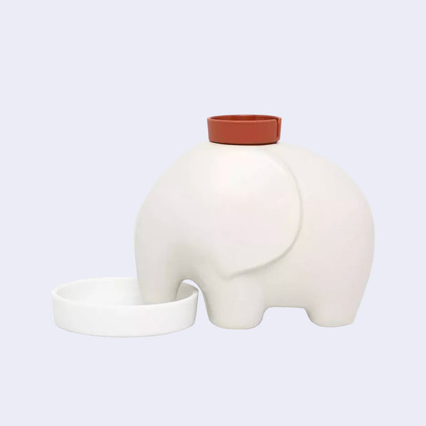 A smooth, white ceramic modern looking elephant with a pot on its head, and its trunk in a shallow white bowl.