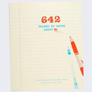 Book cover, cream colored with a vertical red line and many horizontal blue lines, like a sheet of notebook paper. "642 Things to Write About Me" is written in stylized font in upper center. An illustration of a red and blue pen are in the bottom right.