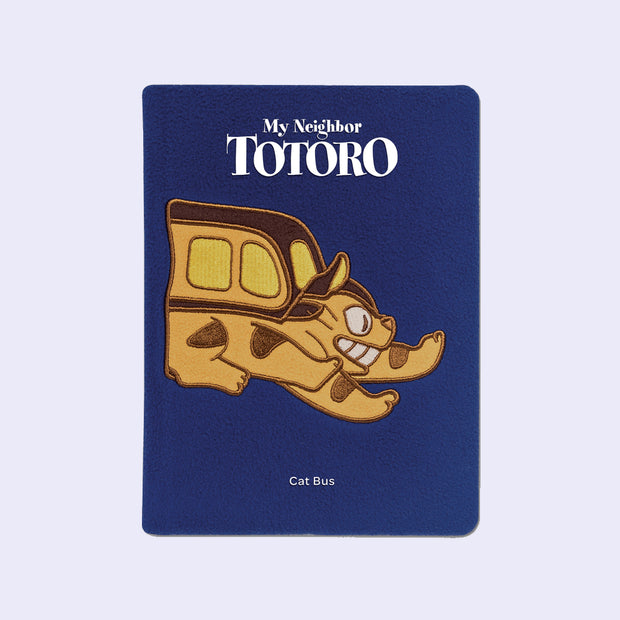 Navy blue plush surface journal, with an embroidery of Catbus from My Neighbor Totoro, only half of its body is showing as it is mid stride. "My Neighbor Totoro" is written on the top of the plastic sleeve with "Cat Bus" written underneath.