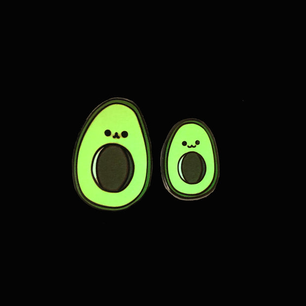 Two enamel pins of illustrated avocados with smiling faces, one is slightly smaller than the other, both are glowing in the dark.