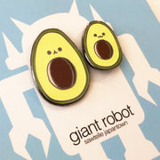 Close up of two enamel pins of illustrated avocados with smiling faces, one is slightly smaller than the other.