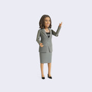 Action figure of politician Kamala Harris, wearing a gray blazer and matching skirt with black heels and a double pearl necklace. She smiles warmly and has one articulate arm raised.