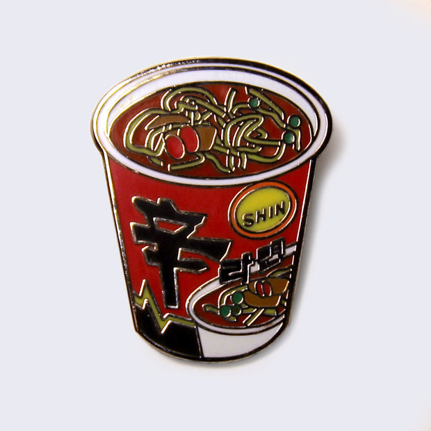 Enamel pin of a red Shin ramen cup packaging, noodles are floating in the cup and there is Japanese script along the side of the cup.