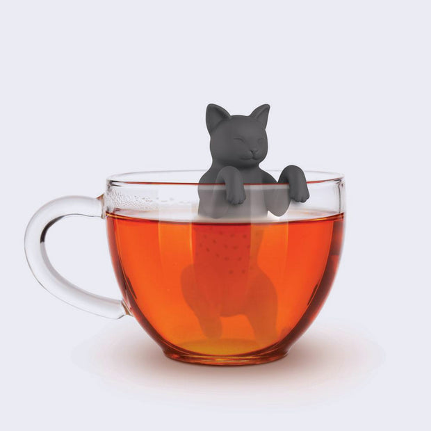 A simple expression gray rubber cat is positioned partly into a clear mug with tea inside of it.