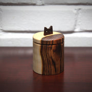 Cylindric wooden box, with a natural wood grain of light wood and darker exterior wood. As a handle for the top is a simplified cat head, with cut out eyes and a small nose.