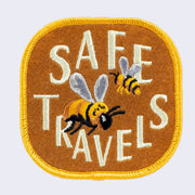 Light chestnut color embroidered patch with gold border. 2 honeybees fly with "Safe Travels" written in stylized all caps cream color font.