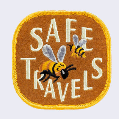 Light chestnut color embroidered patch with gold border. 2 honeybees fly with "Safe Travels" written in stylized all caps cream color font.