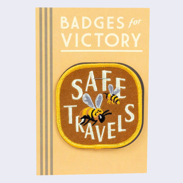 Light chestnut color embroidered patch with gold border. 2 honeybees fly with "Safe Travels" written in stylized all caps cream color font. Patch is on a light yellow backing card.