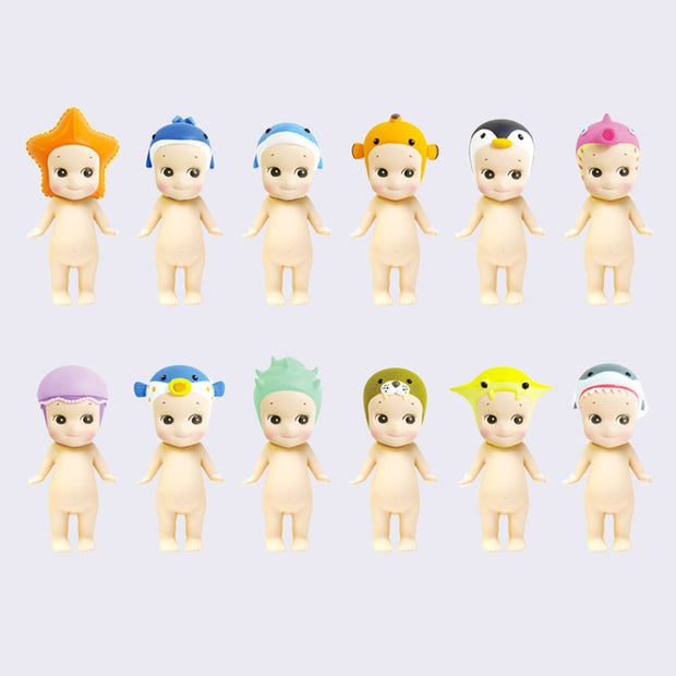 12 vinyl Kewpie babies, standing nude with different marine themed creatures atop their heads like hood hats. Options are: starfish, whale, dolphin, clownfish, penguin, seahorse, jellyfish, blowfish, shell, otter, manta and shark.