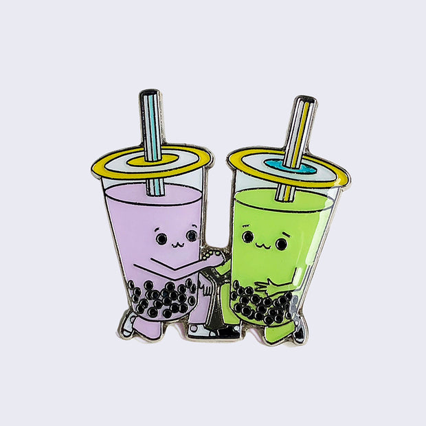 Enamel pin of a purple taro boba cup and a green matcha boba cup kneeling and clasping hands.