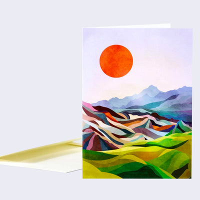 Greeting card with full bleed illustration of a scene of an open mountain range, comprising of many triangular shapes and colors. Overhead a large orange sun hangs against a light pink sky.